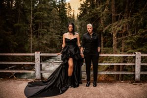 A couple poses on a wooden bridge in a forest at sunset, with the woman wearing a dramatic black flowing gown.