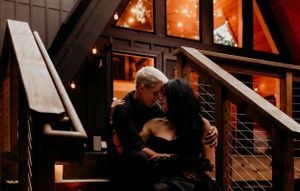A cozy scene of a couple embracing on steps outside a wooden cabin with warm glowing windows at night.