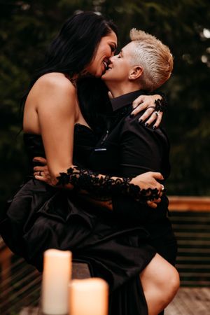 Romantic couple in black outfits sharing an intimate moment on a deck with candlelight at dusk.