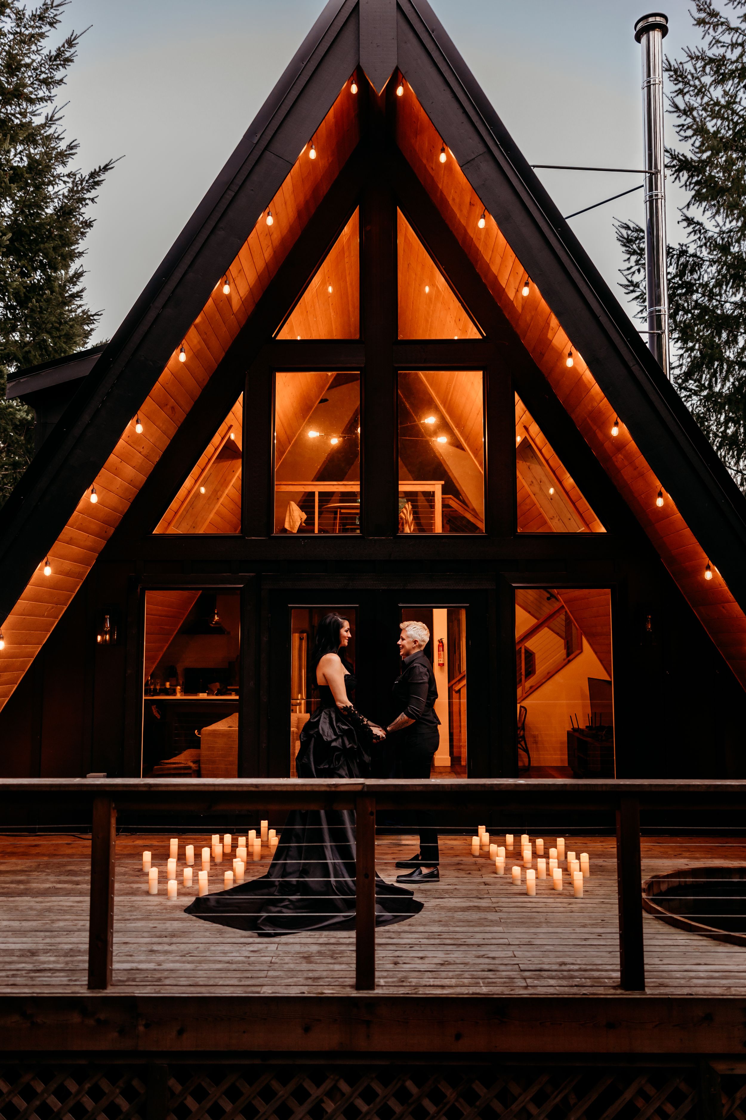 A-frame cabin lit with warm string lights at dusk with silhouettes visible through large triangular windows and candles on the deck.