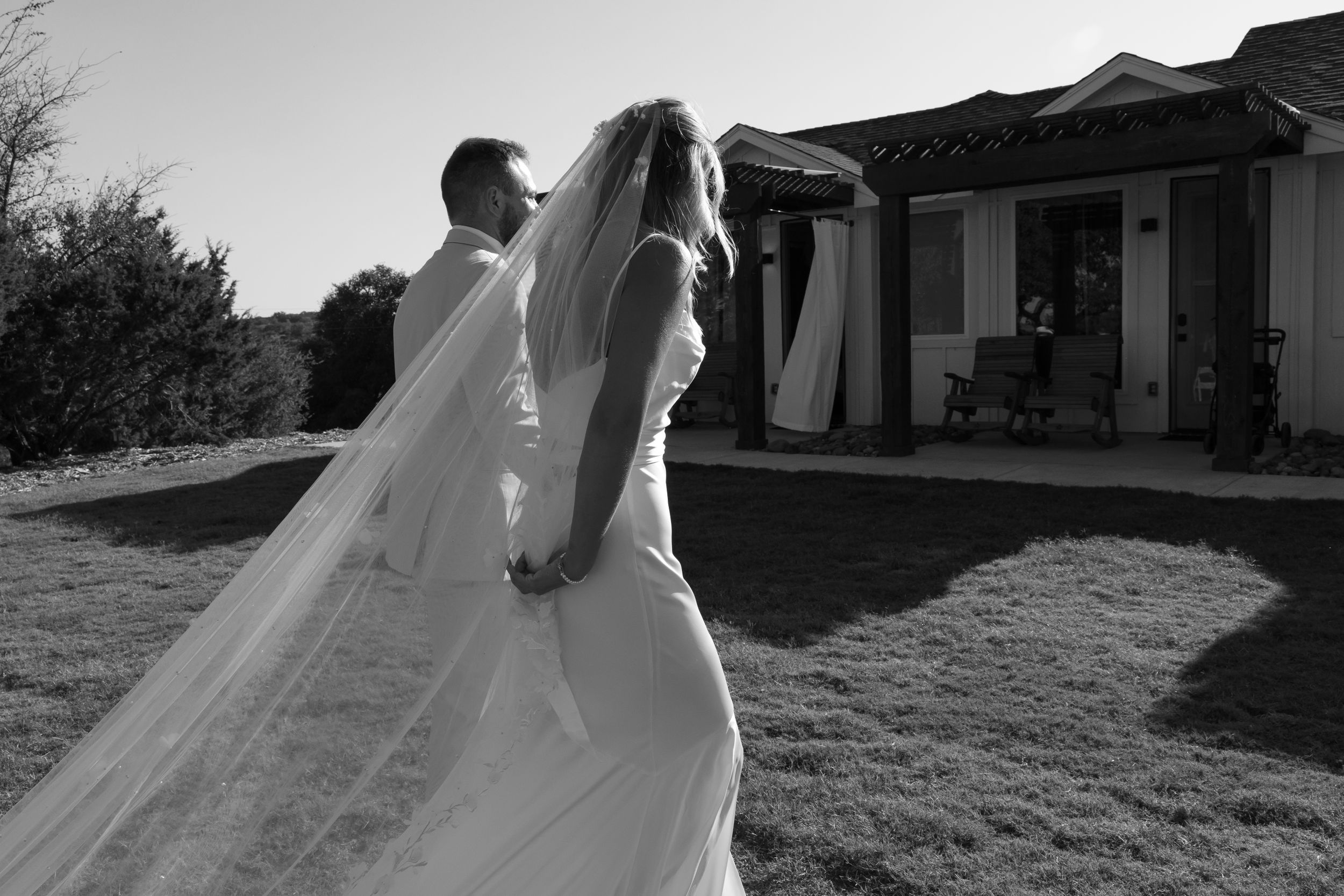 Newlyweds walk on grassy lawn outside venue with long flowing white veil trailing behind in black and white.