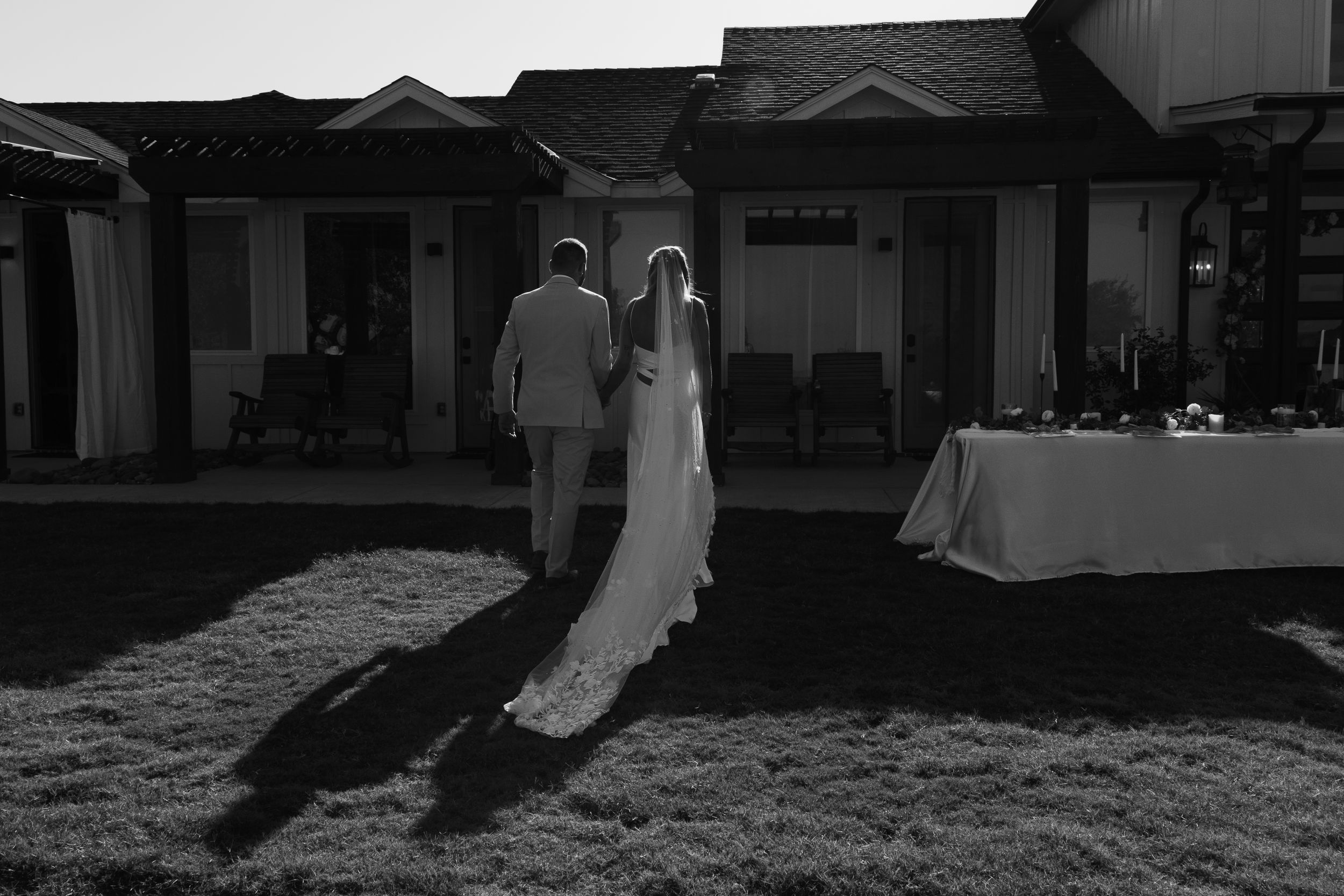 A black and white wedding scene shows a couple casting long shadows on a lawn during an outdoor reception.