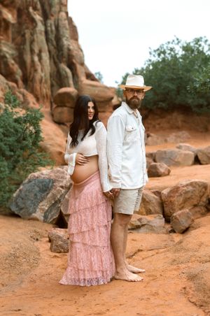 A couple poses on a red rock trail surrounded by desert cliffs, with her wearing a pink layered skirt and him in a white shirt.