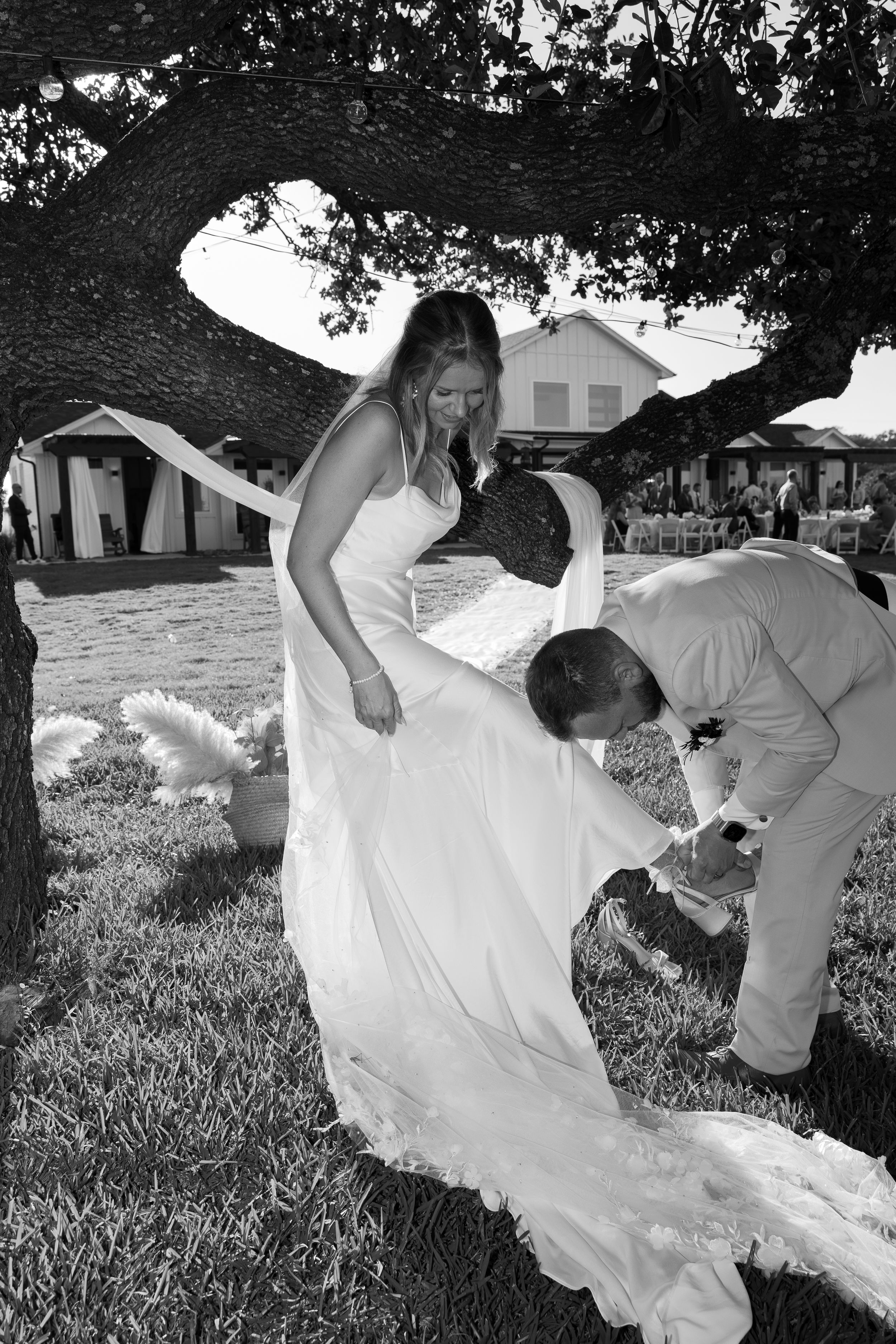 A wedding couple shares a candid moment under a tree branch as they adjust attire outdoors in black and white.