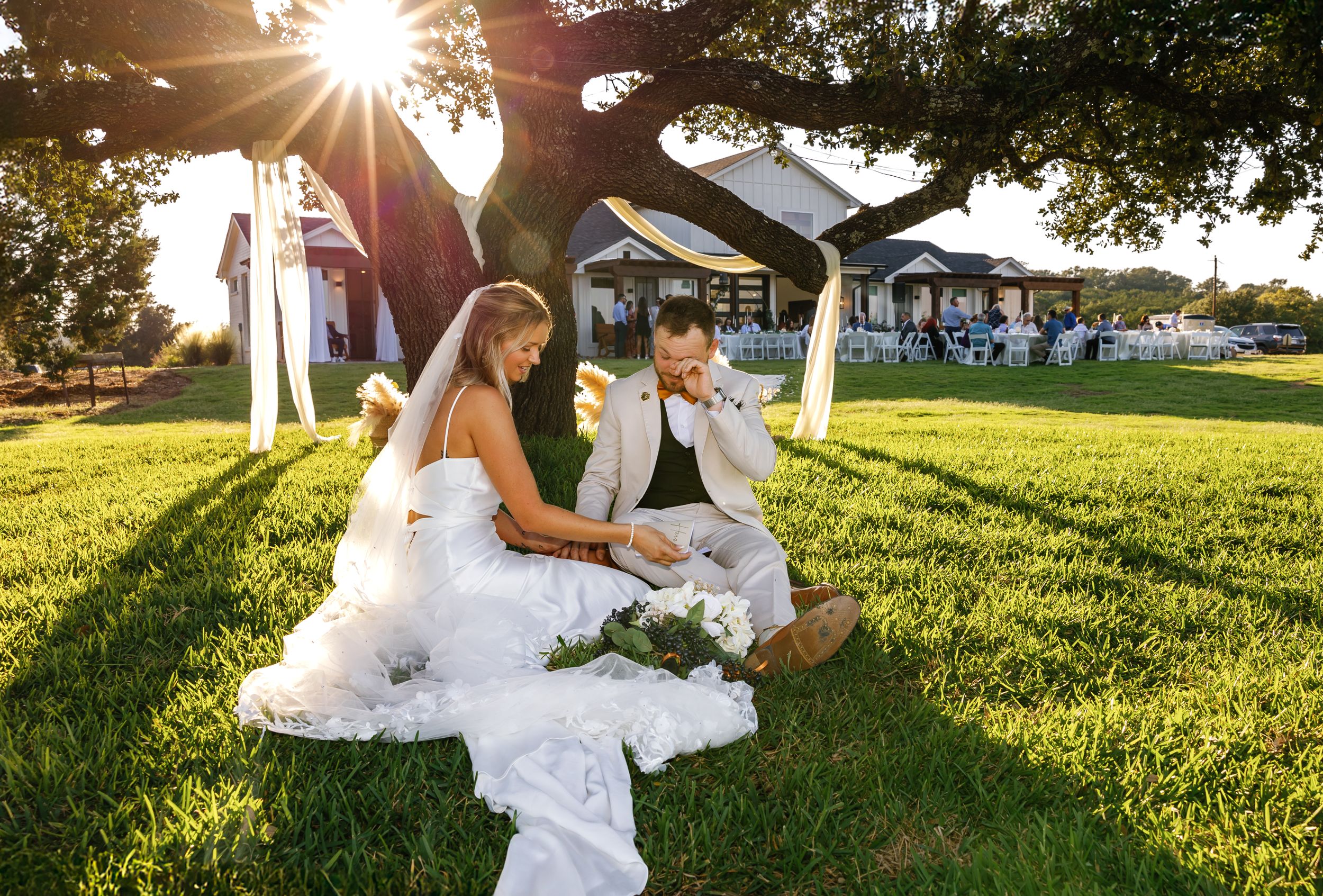 Two people in wedding attire sit together on grass during golden hour sunset at an outdoor venue.
