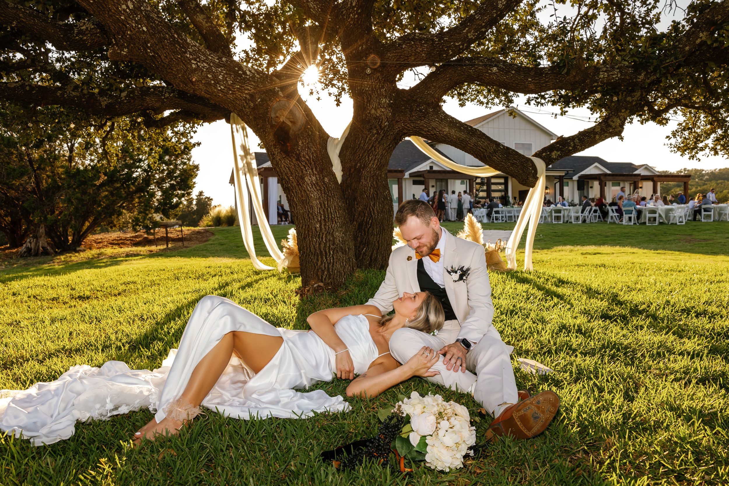 Newlyweds embrace in romantic pose under large oak tree at sunset during outdoor wedding celebration.
