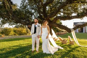 A couple in wedding attire poses under a large tree at sunset during an outdoor ceremony.