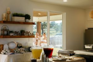 Sunlit kitchen countertop with floating wooden shelves, drinks in glass pitchers, and decorative items against a window view.