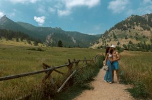 Two people walk along a scenic mountain trail with rustic wooden fence and rolling foothills in Colorado wilderness.