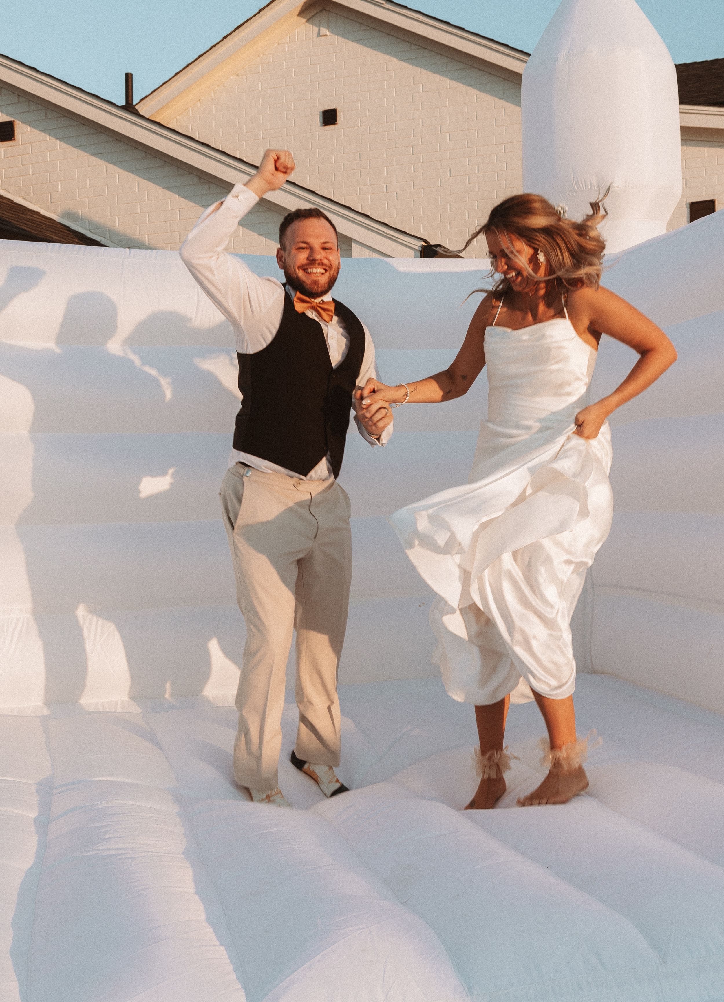 Happy couple dancing and jumping on a white bounce house at an outdoor wedding celebration during sunset.