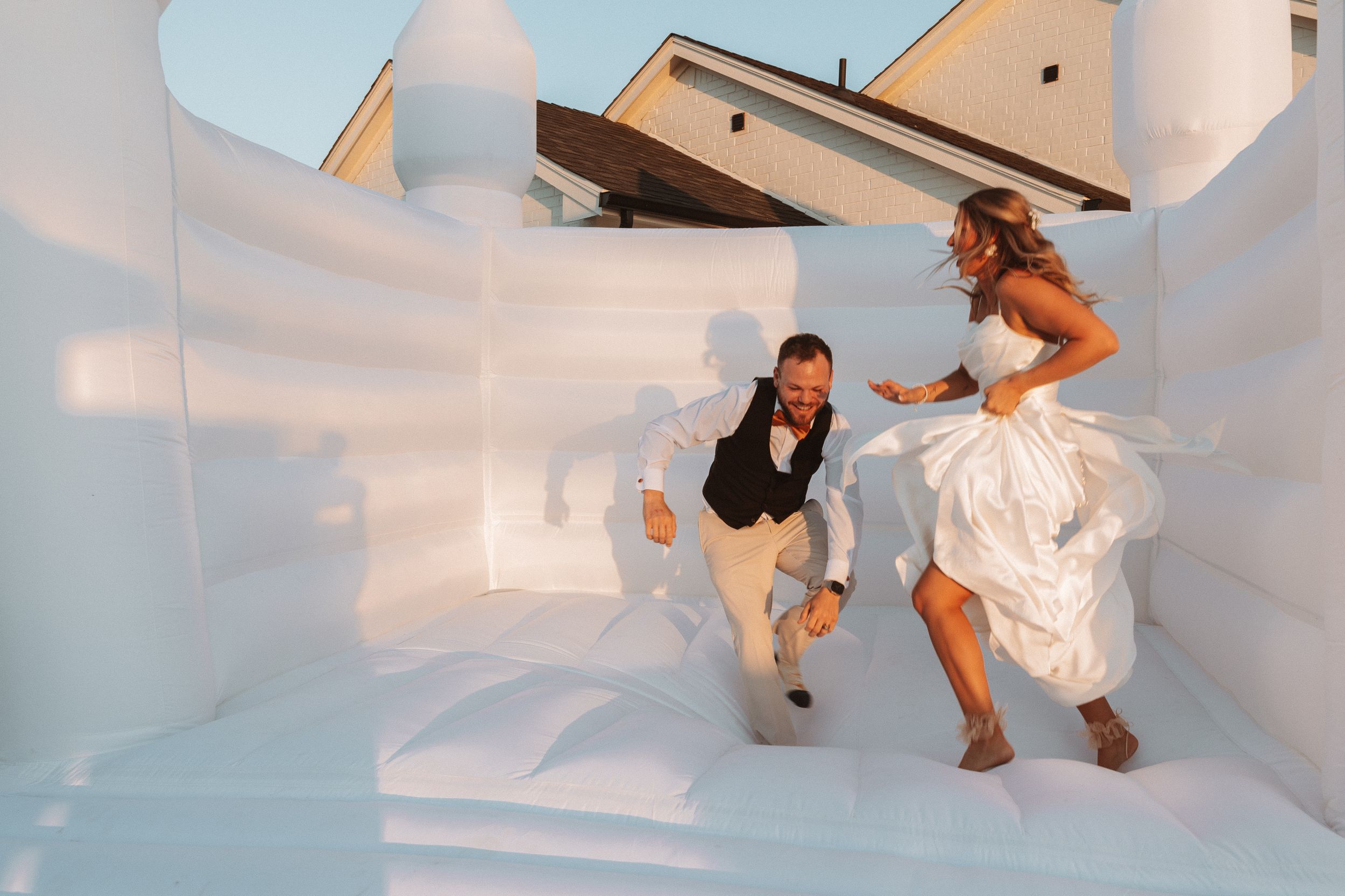 Couple joyfully jumping inside an all-white bouncy castle or inflatable structure at wedding celebration.