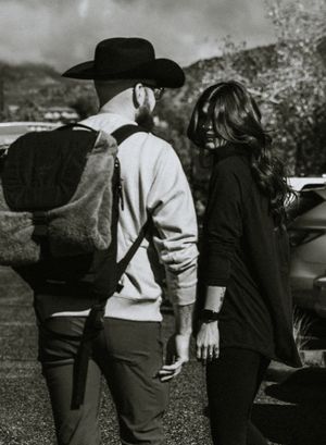 A black and white photo of two people walking outdoors, one wearing a cowboy hat and backpack.