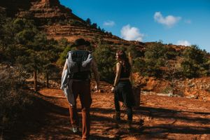 Hikers walking along a scenic red dirt trail surrounded by desert vegetation on a sunny day.