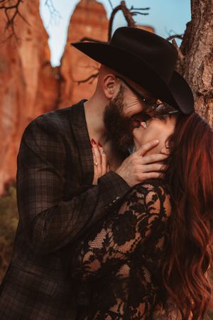 A romantic couple sharing a passionate kiss against red rock formations while he wears a black cowboy hat and suit.