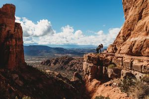 A hiker on a narrow mountain trail overlooking a vast desert canyon with red sandstone cliffs and blue skies.