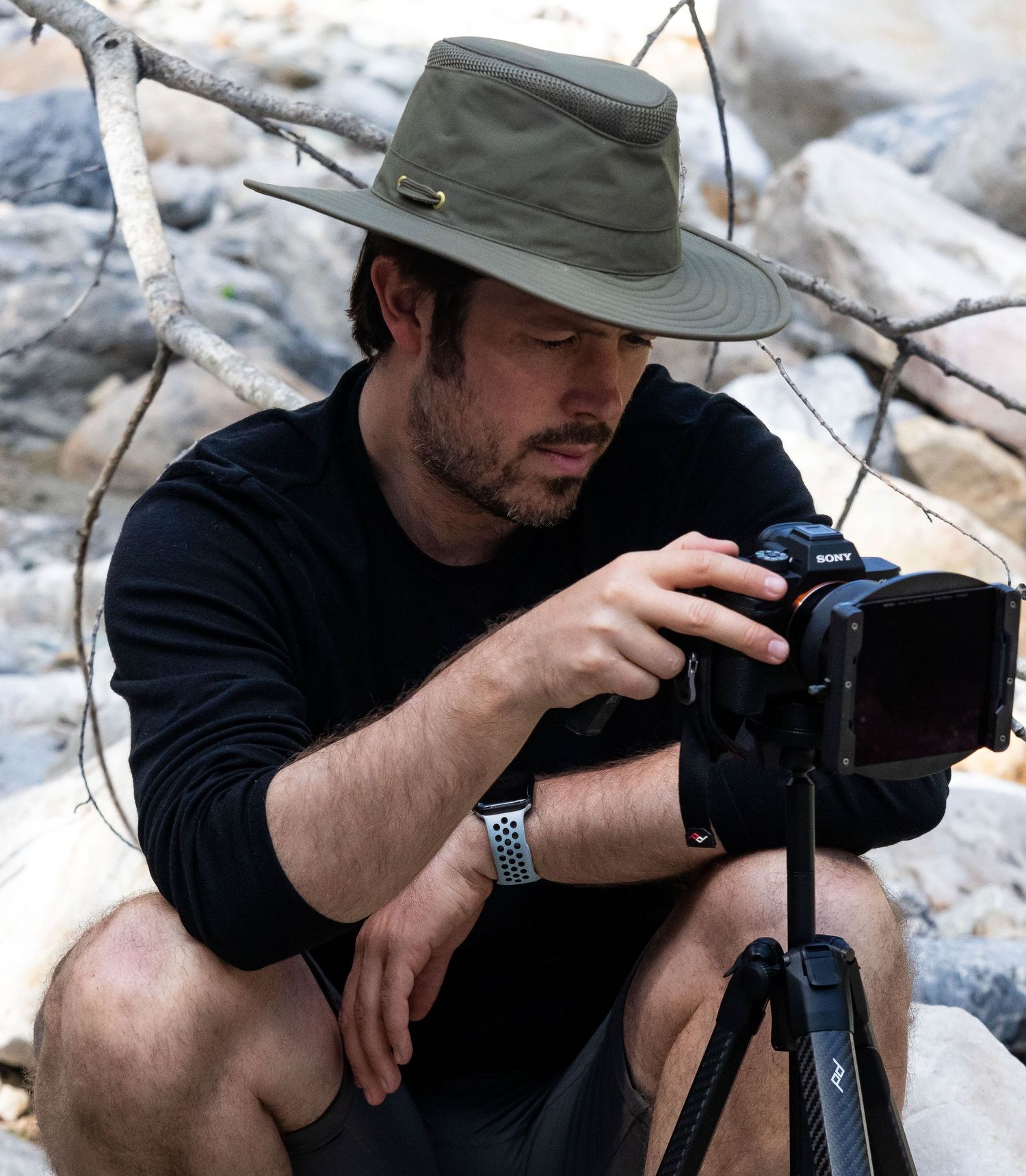 Photographer Evan Will wearing green hat and black shirt adjusts camera on tripod in rocky outdoor setting by a river in the mountains