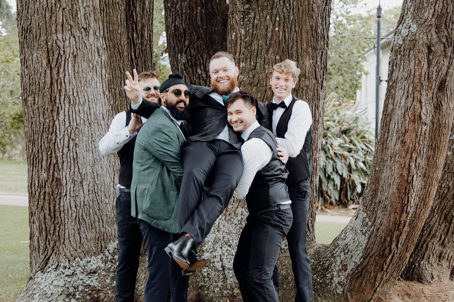 The groom and the guys pose playfully between large tree trunks in an outdoor setting, captured by Apertured Media Auckland Wedding Photogr