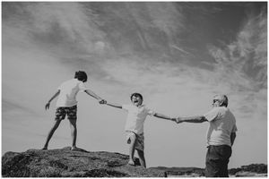 Three people forming a human chain while balancing on coastal rocks against the sky.