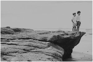 Two figures standing on a dramatic rocky cliff overlooking the ocean in black and white.