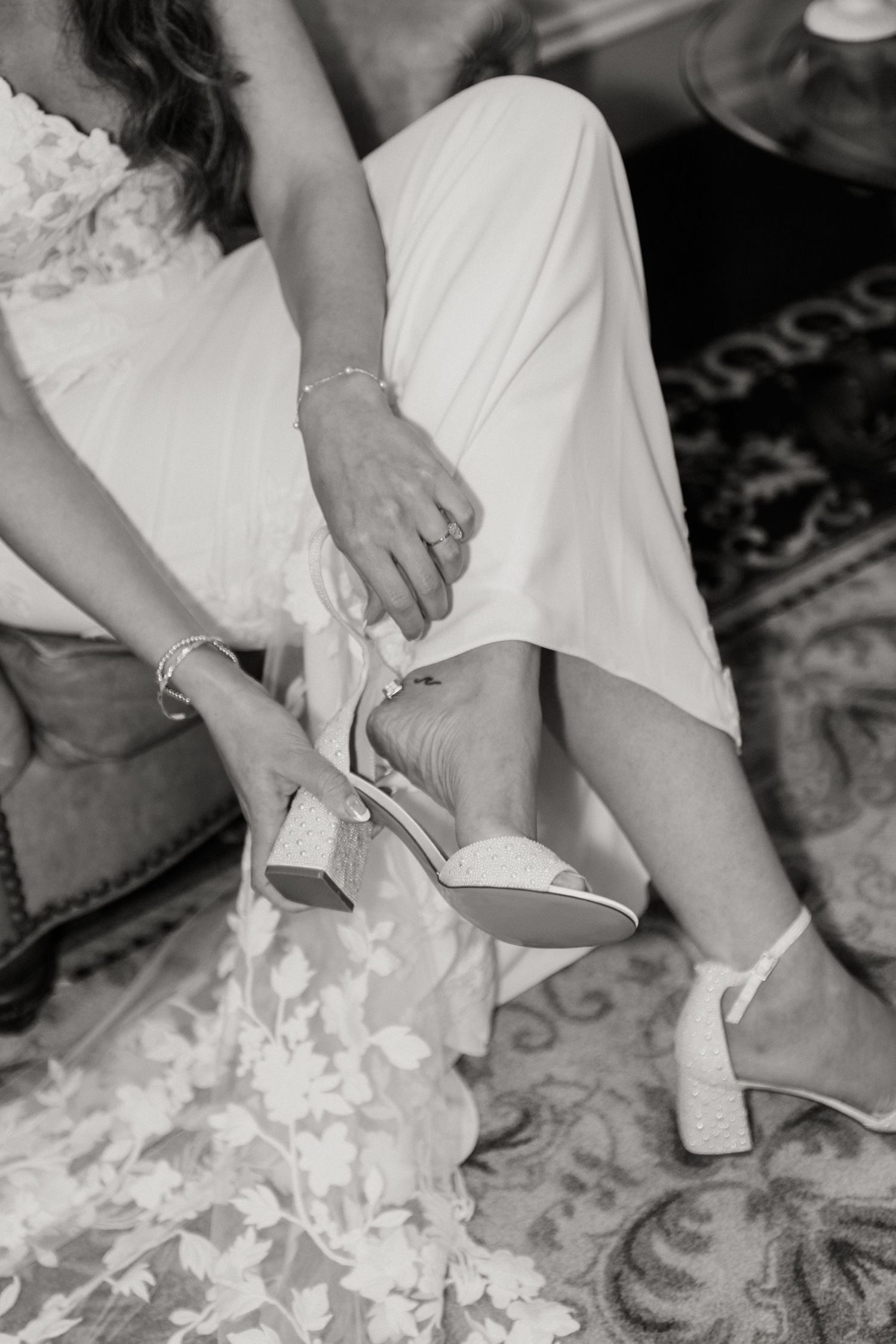 Black and white photo of bride adjusting wedding shoe while sitting.