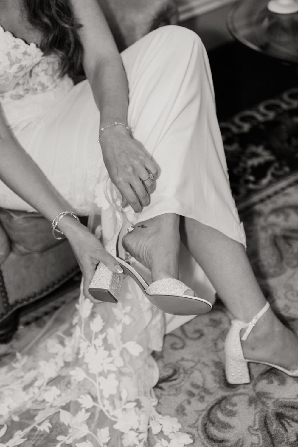 Black and white photo of bride adjusting wedding shoe while sitting.