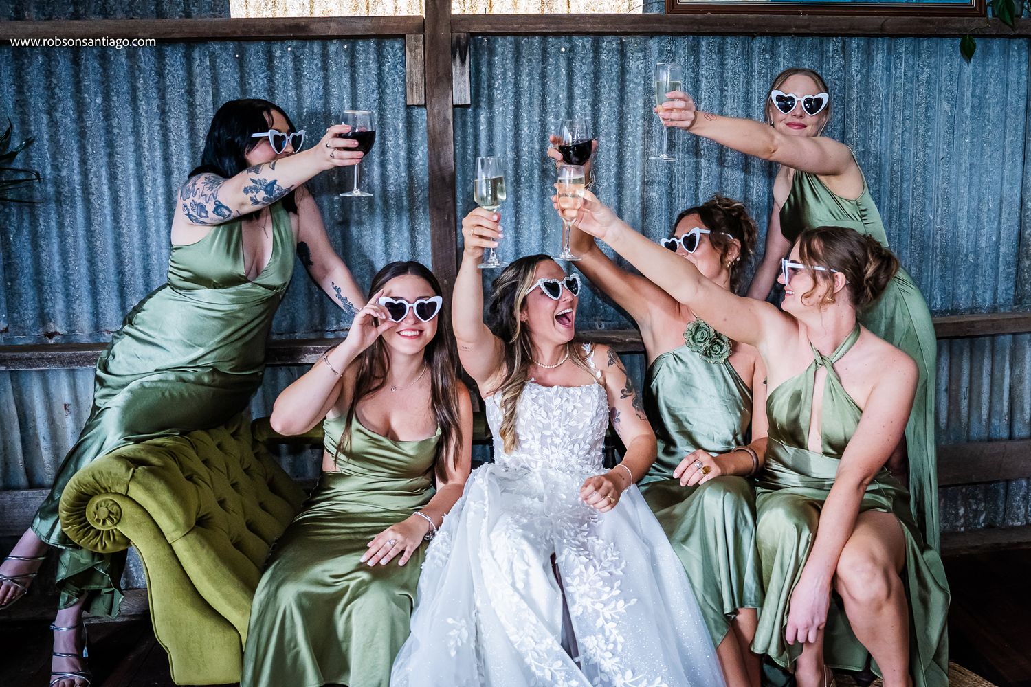 Bridal party in sage green dresses toasting and posing playfully against a rustic blue metal wall.