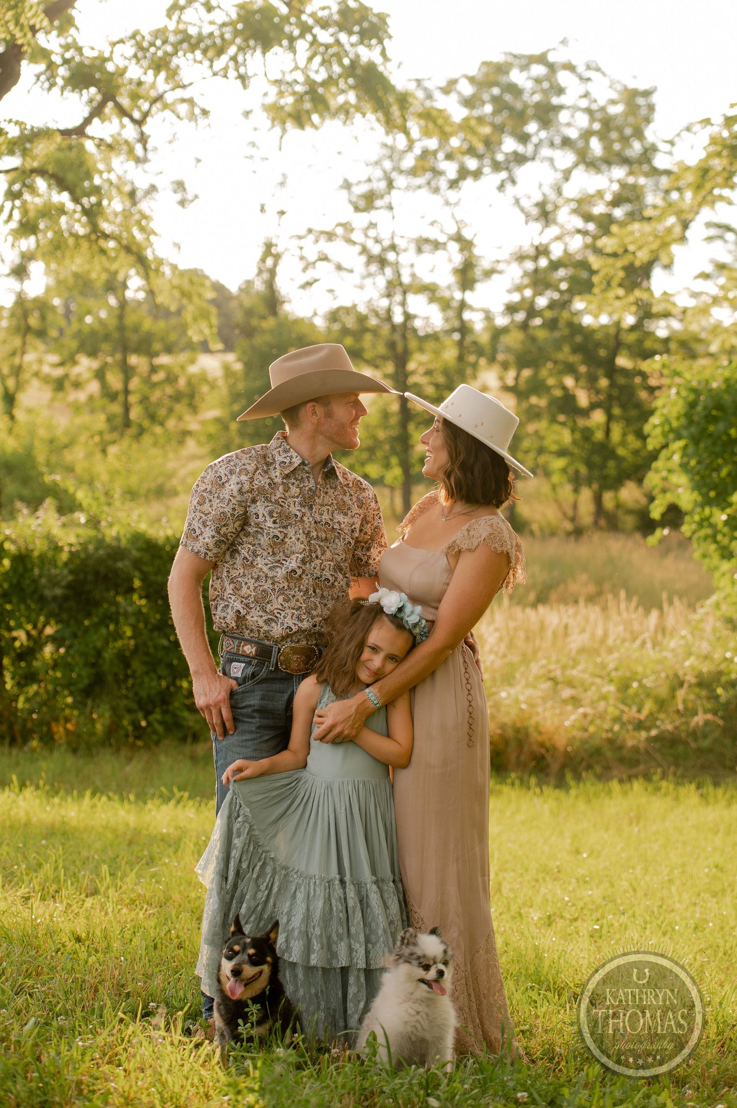 Warm sunset family portrait with two small white dogs in a rustic outdoor setting.