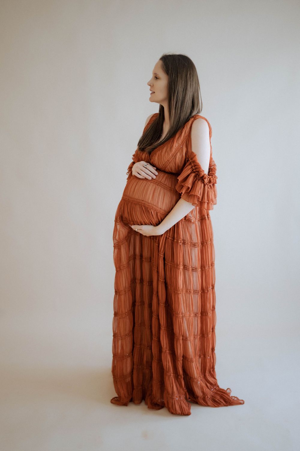 A pregnant person in profile wearing a flowing rust-colored tiered maxi dress stands against a light gray background.