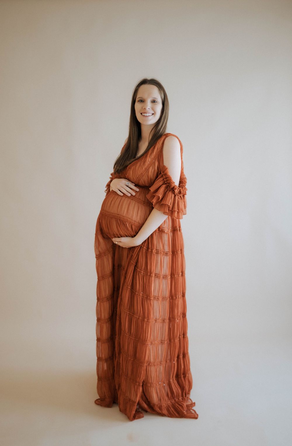 A woman in a flowing rust-colored tiered maternity dress poses against a neutral backdrop during a pregnancy photoshoot.