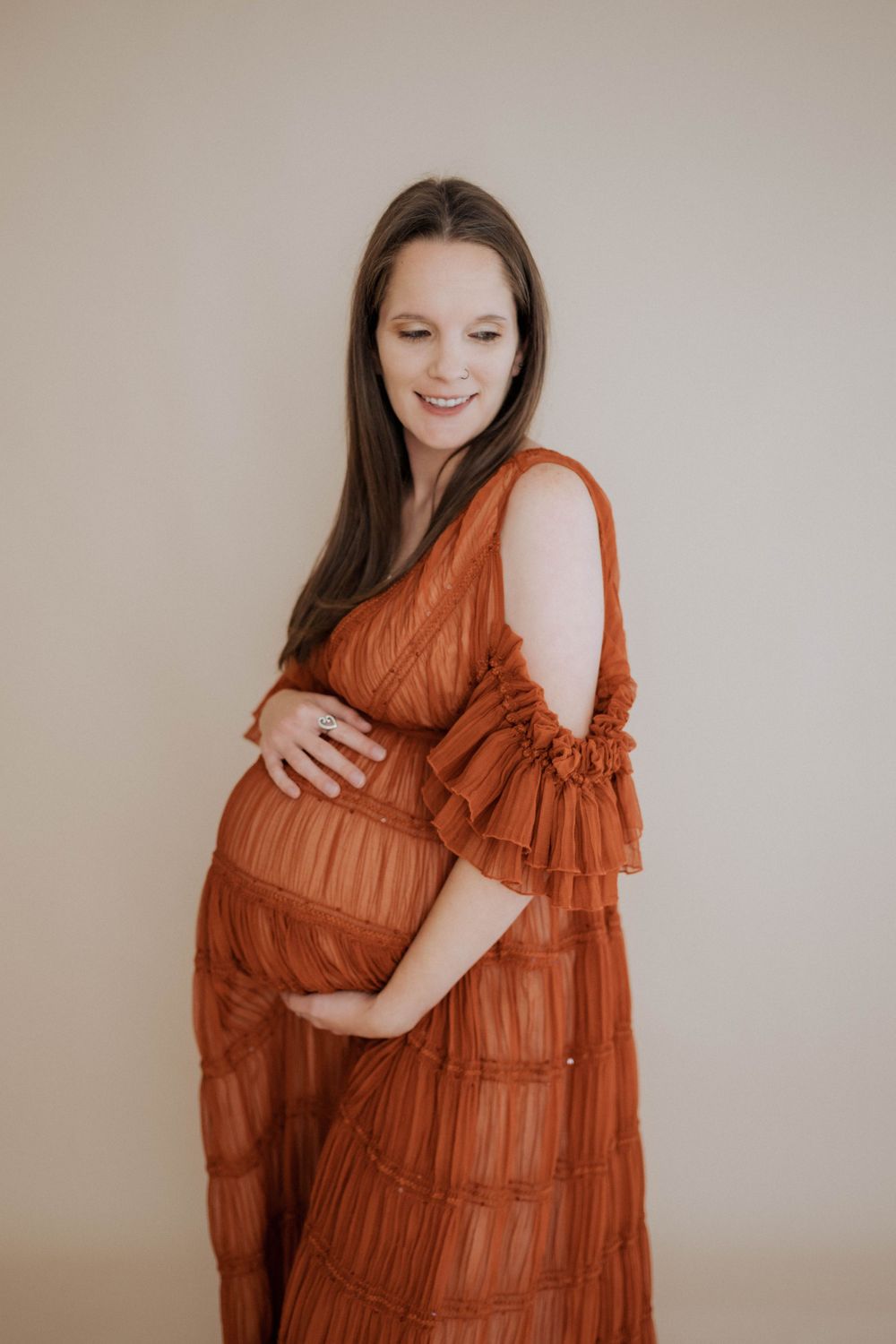 A maternity photo shows a long-haired person in a rust-colored tiered maxi dress with ruffled sleeves against a neutral background.