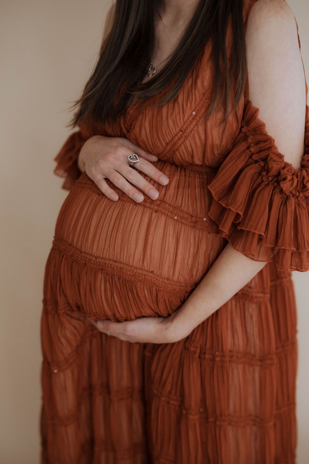 Close-up view of a pregnant belly in a rust-colored tiered dress with ruffled detail and hands gently cradling the bump.