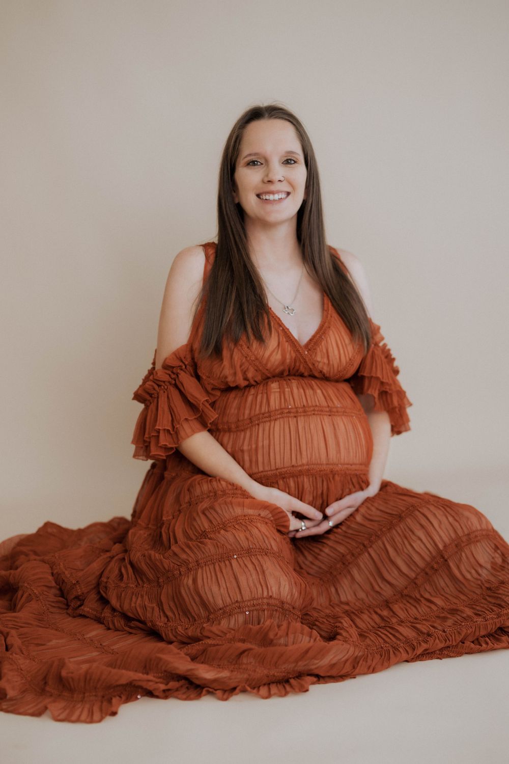 Expectant woman in flowing rust-colored maxi dress with ruffled details poses for maternity photo against neutral backdrop.