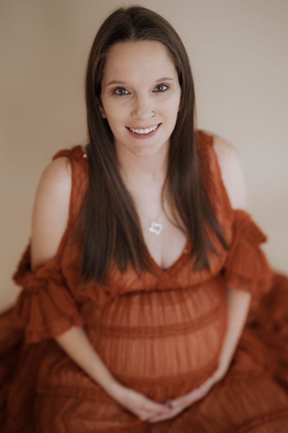 A maternity portrait of someone wearing a rust-colored flowy dress with ruffled sleeves and a necklace against a beige background.