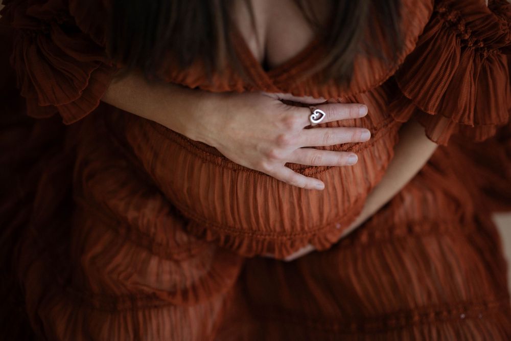 A hand wearing an engagement ring rests on a rust-colored ruffle dress in a moody artistic composition.