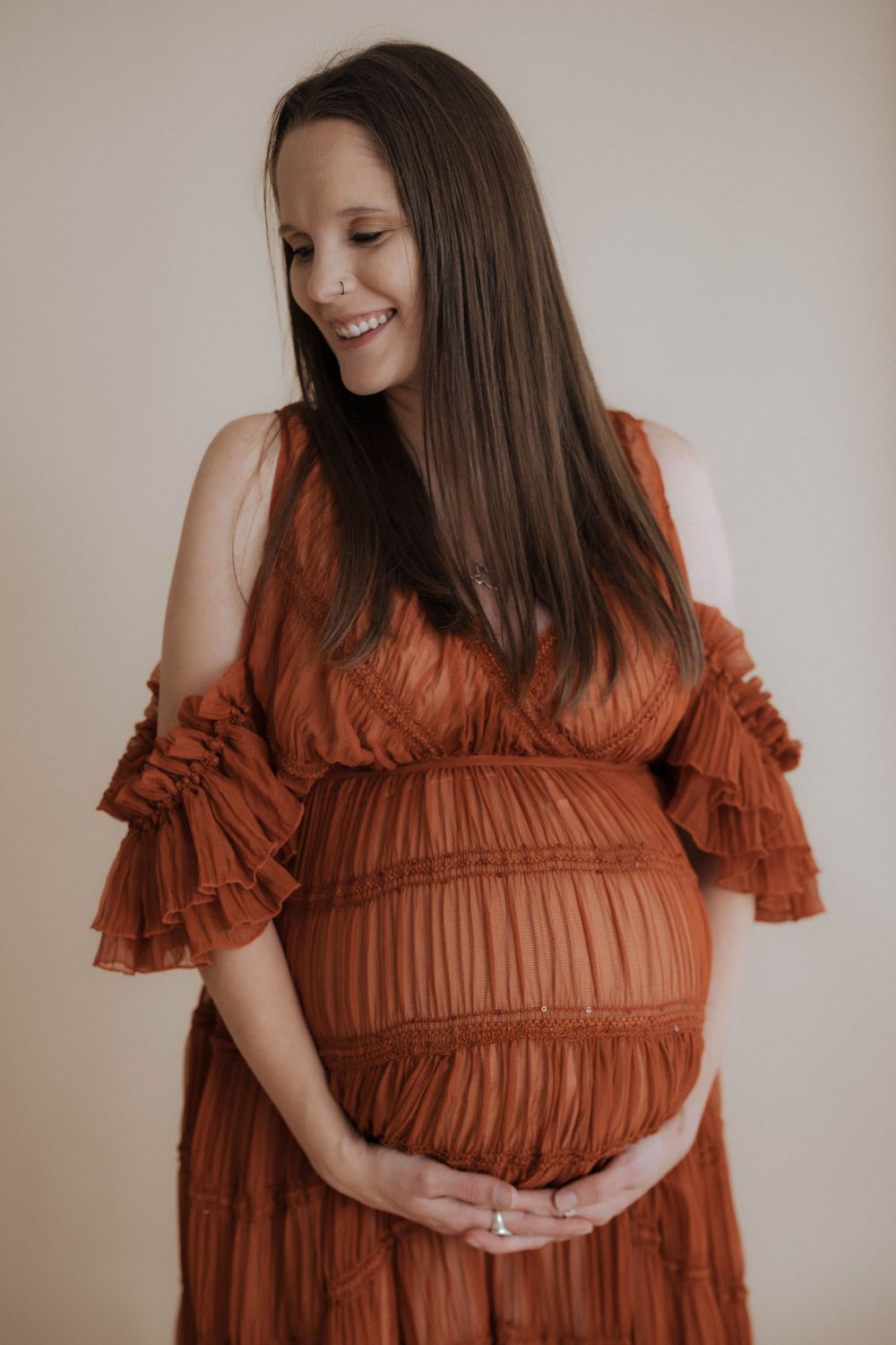 Expectant woman in rust-colored pleated dress with ruffled sleeves poses against neutral wall during maternity photoshoot.