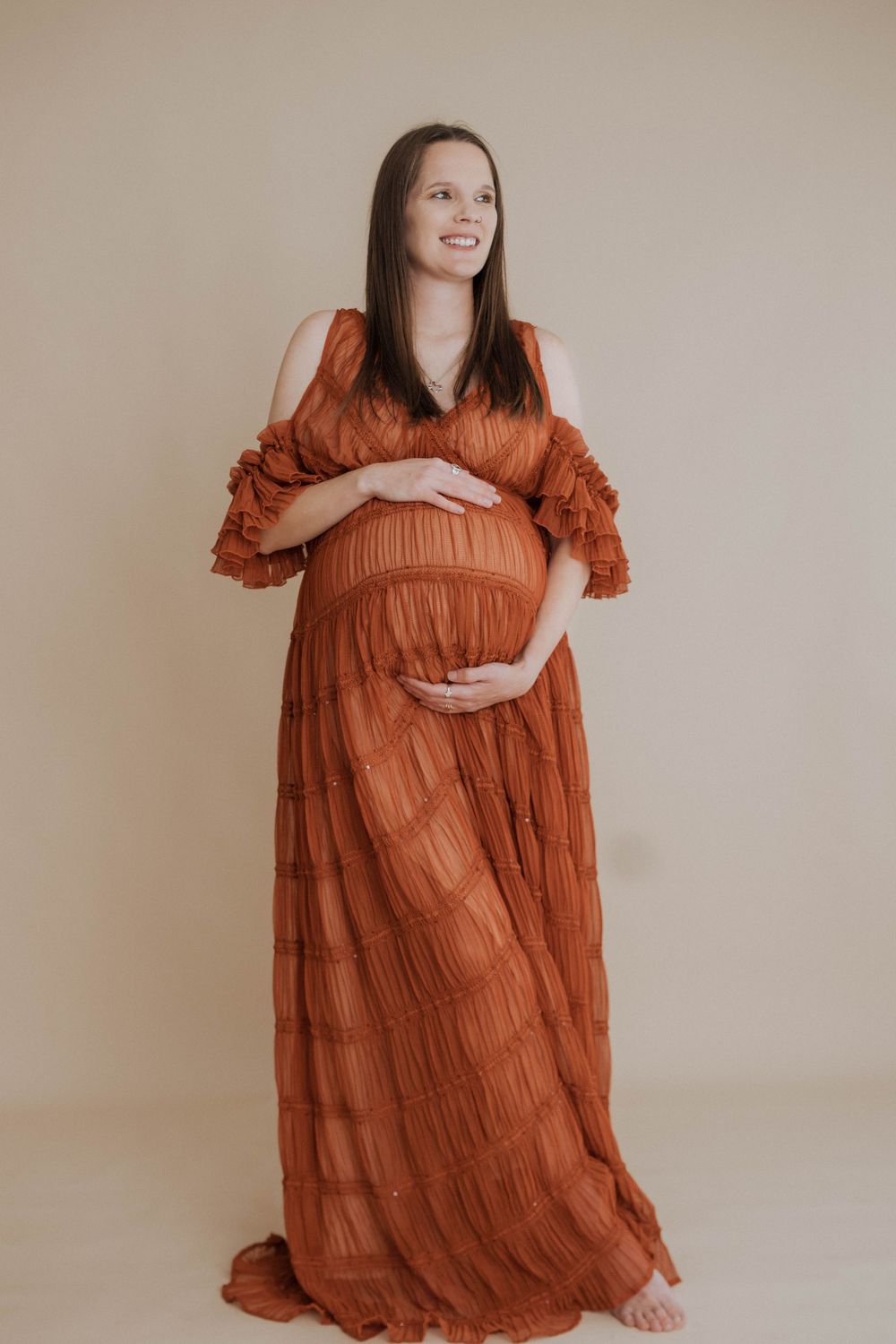 Expectant mother wearing a flowing rust-colored tiered maxi dress poses against neutral backdrop while cradling baby bump.