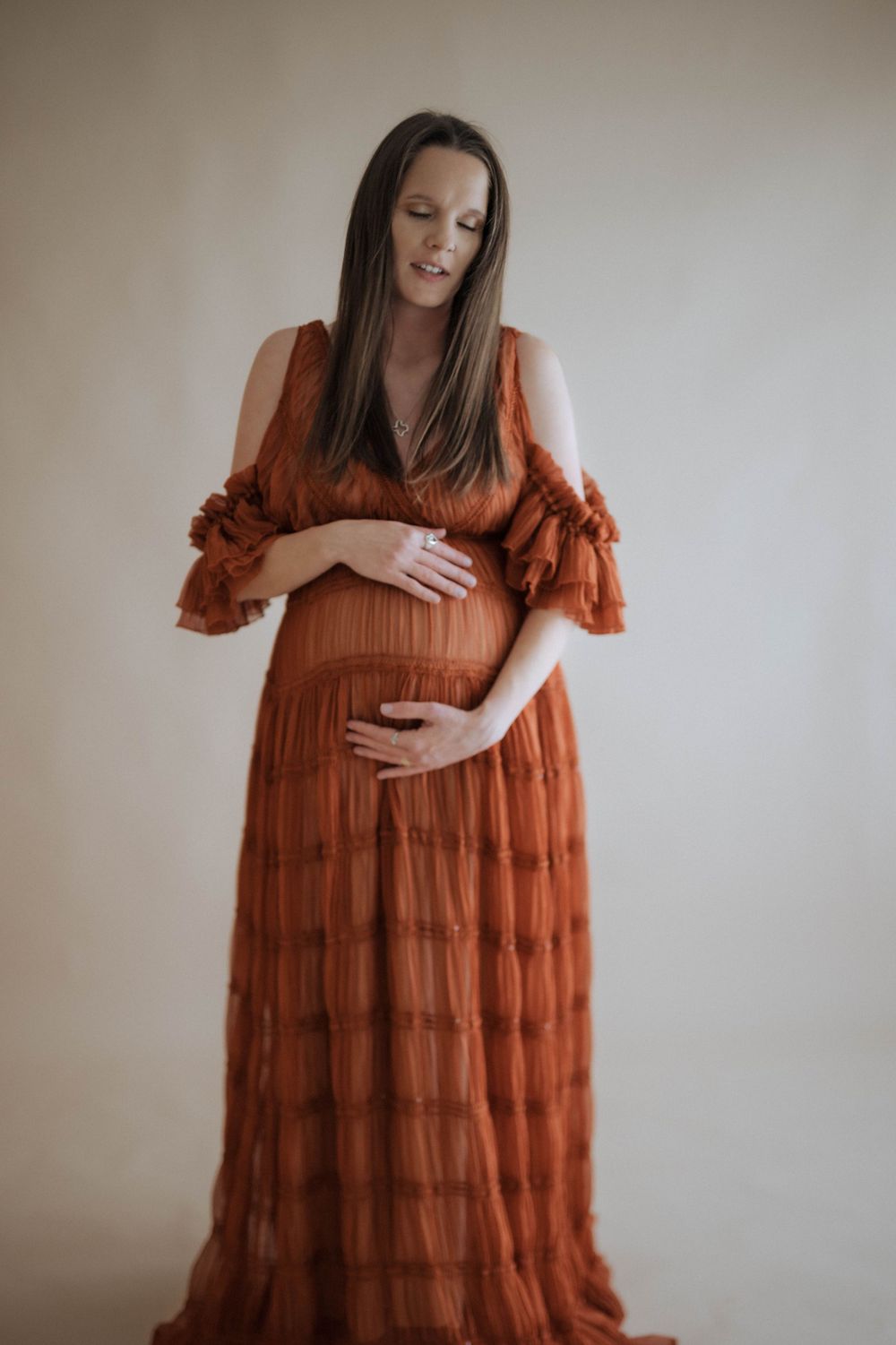 A maternity portrait features a person in a flowing rust-colored dress with ruffle details against a light wall.