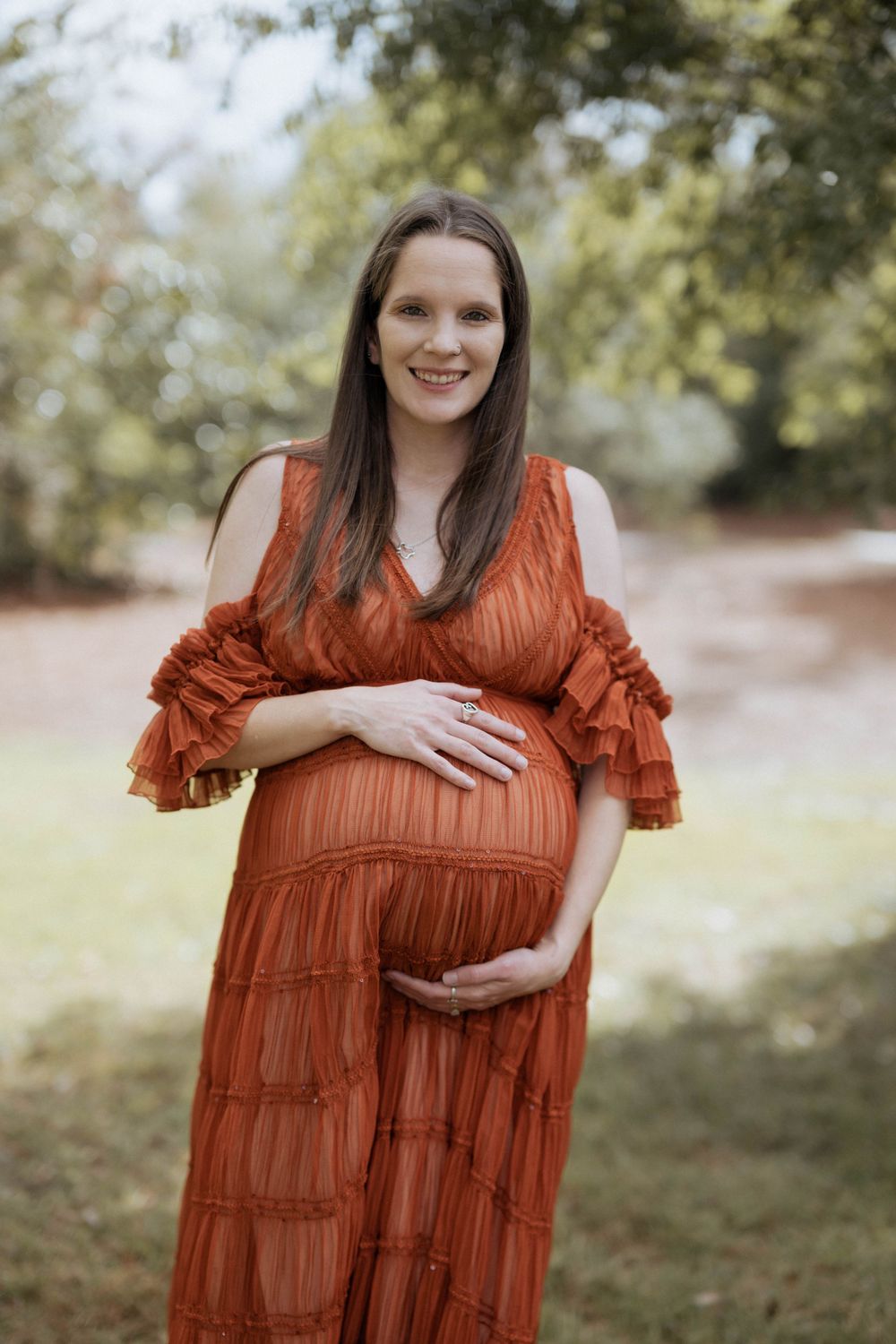A pregnant person in a flowing rust-colored dress with ruffled sleeves poses outdoors on a sunny autumn day.