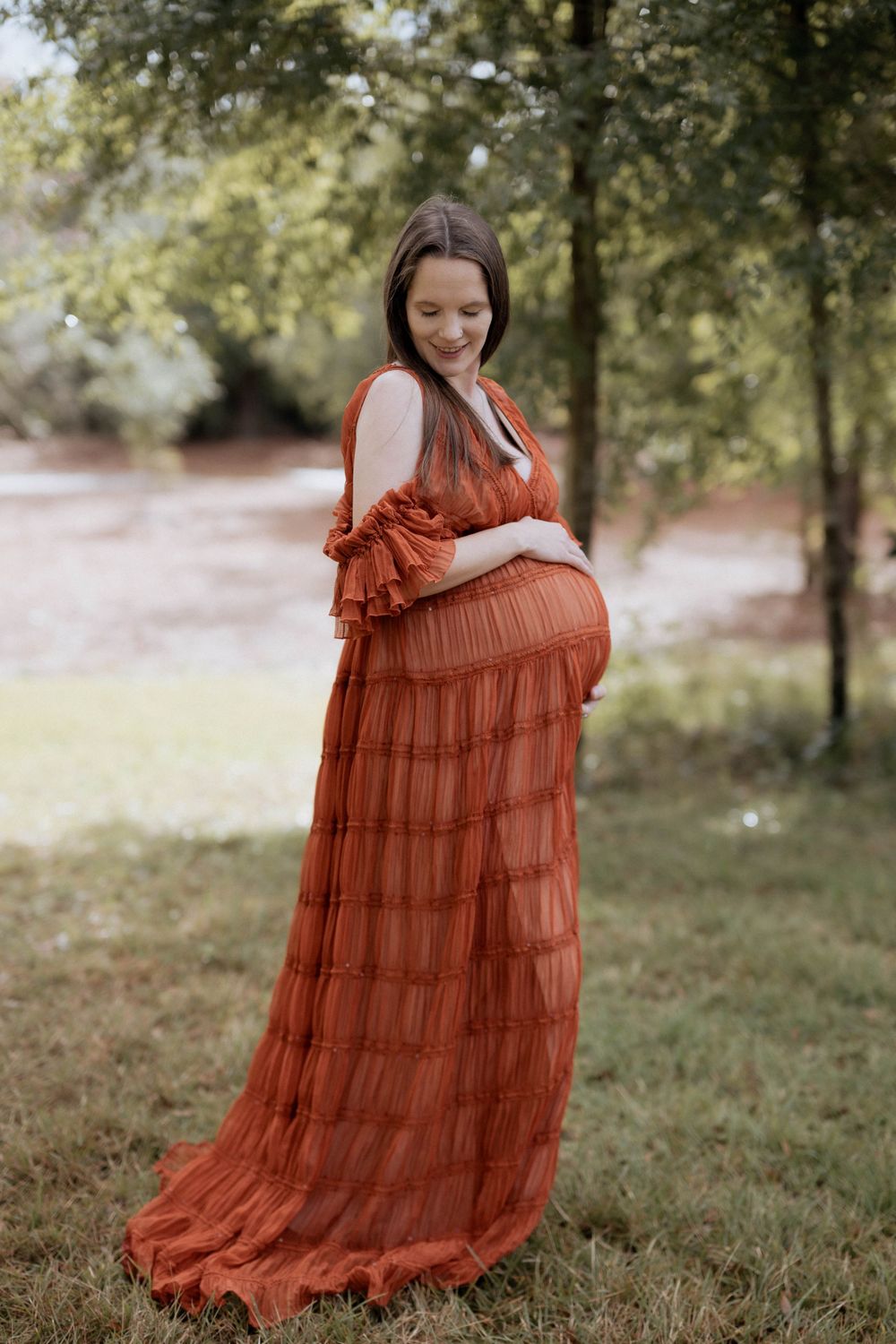 Expectant mother wearing a flowing rust-colored tiered maxi dress poses outdoors in a natural woodland setting.