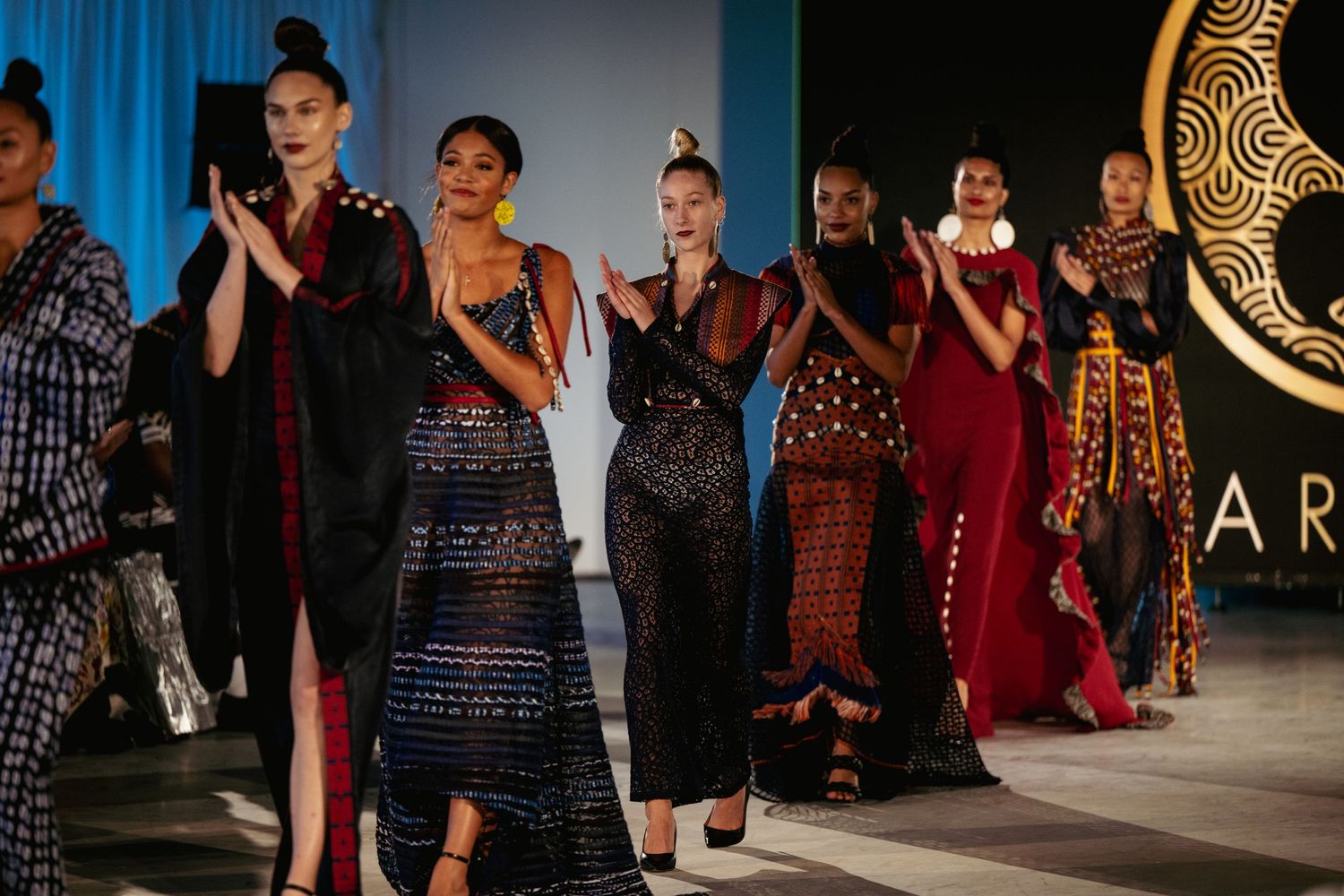 Models walk the runway wearing elegant evening gowns in dark colors at a fashion show.