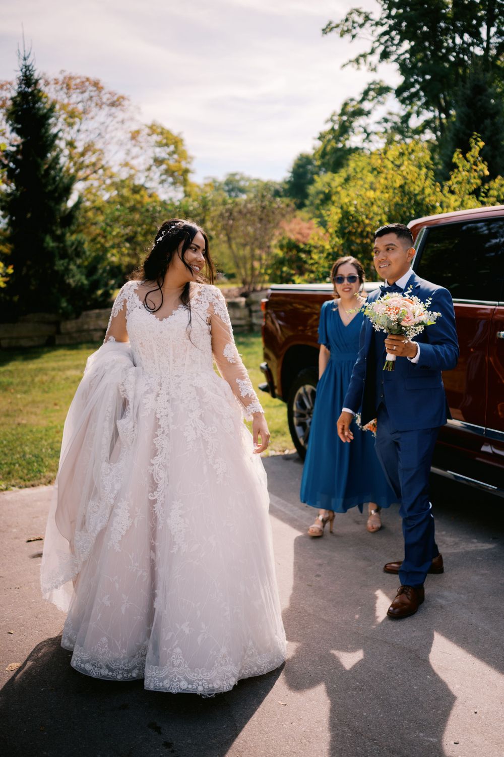 A bride in a lace gown walks near a vintage car while attendants follow behind.