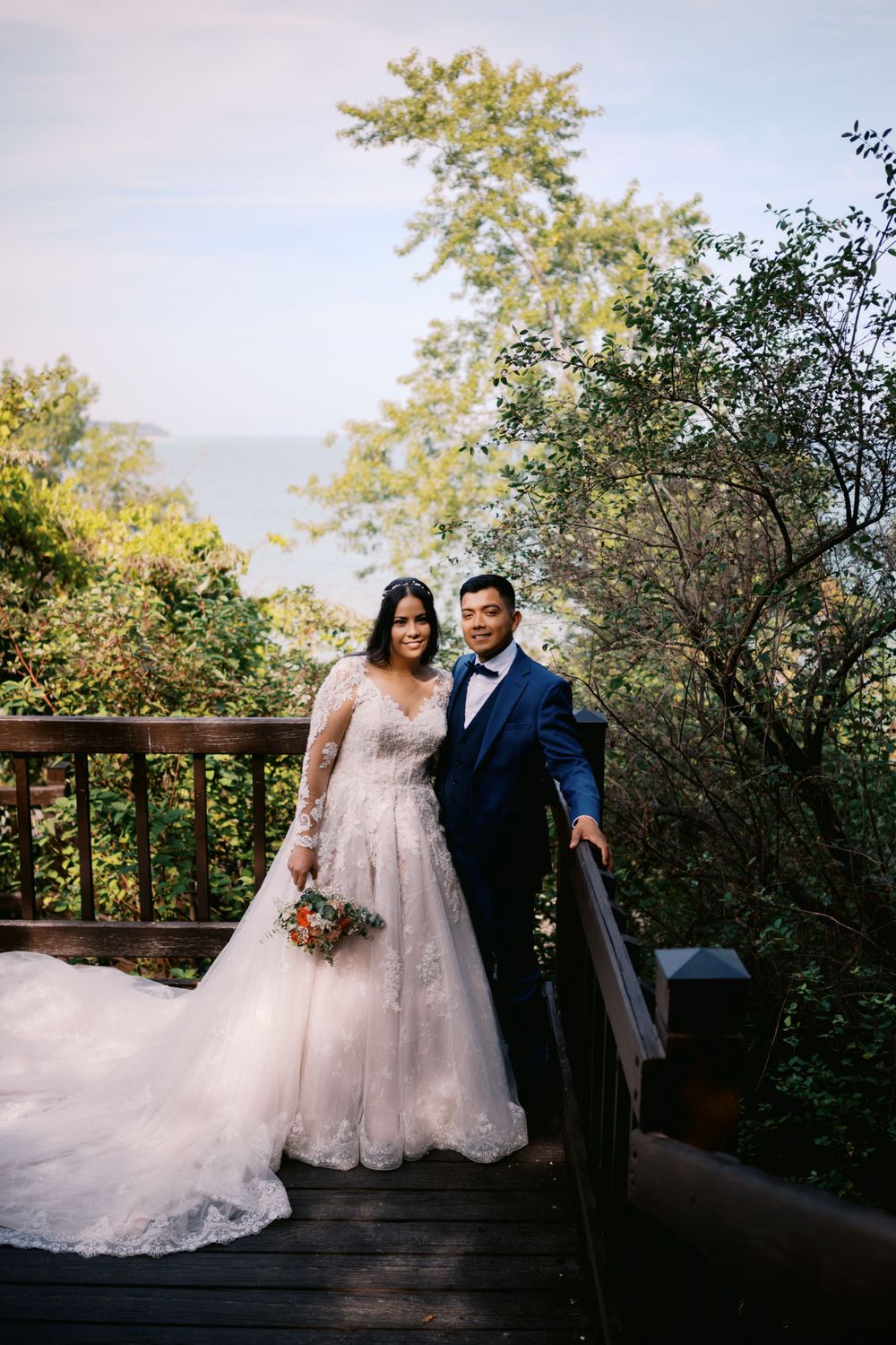 A couple poses on a scenic wooden deck surrounded by lush greenery in wedding attire.