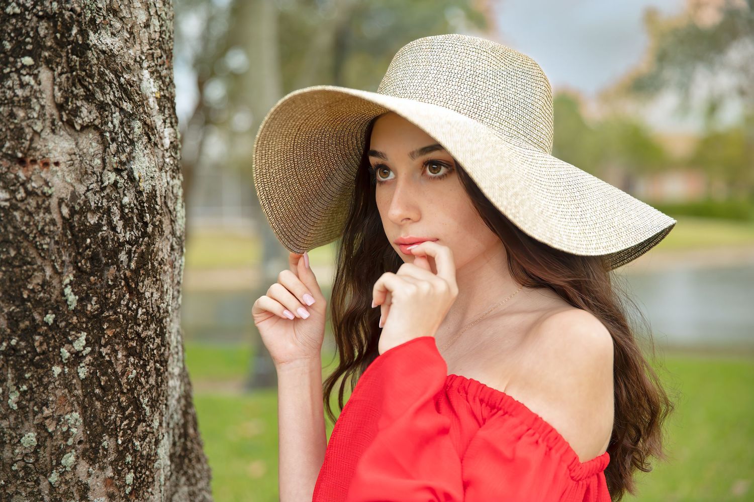 Person in red off-shoulder dress and wide-brim sun hat posing by tree.