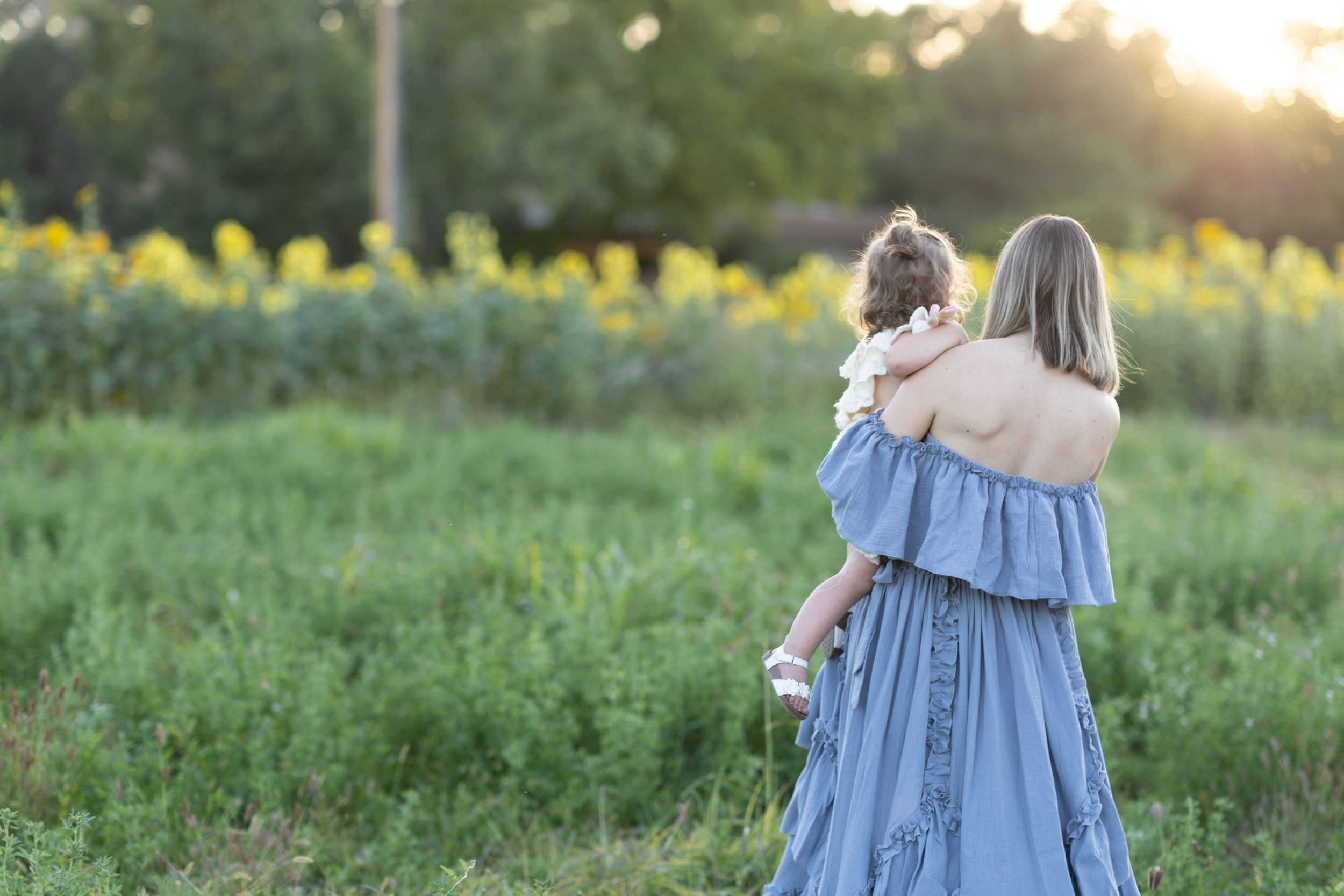 A figure in a flowing blue dress holds a small child while standing in a sunlit field with wildflowers.