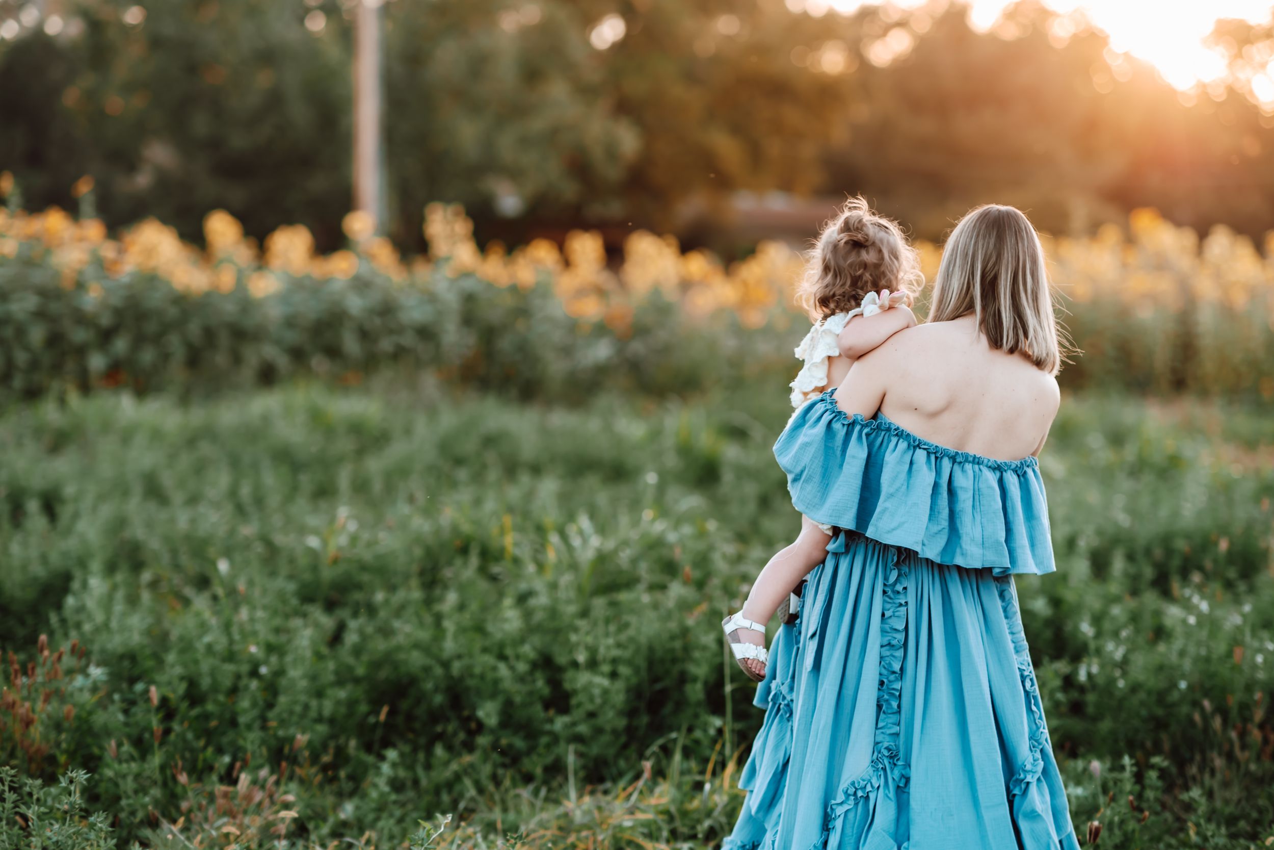 Figure in a flowing turquoise dress holding a child while standing in a sunlit meadow at golden hour.