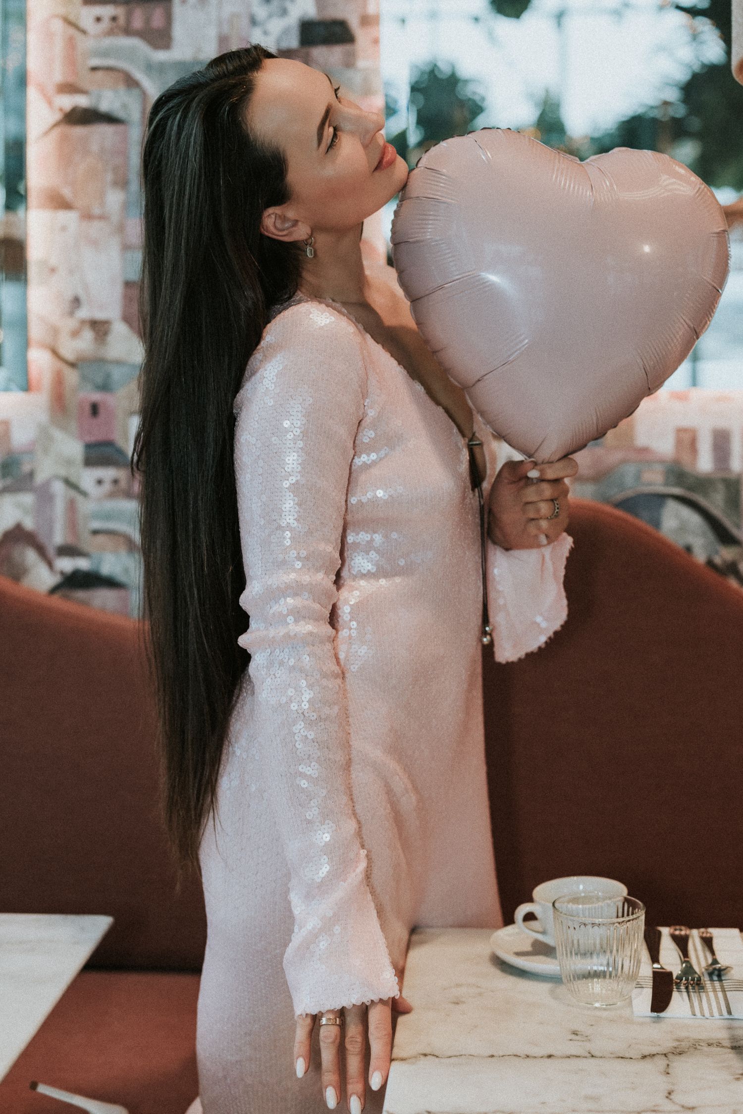 Woman with long dark hair in pink sequin dress holding heart balloon in restaurant setting.