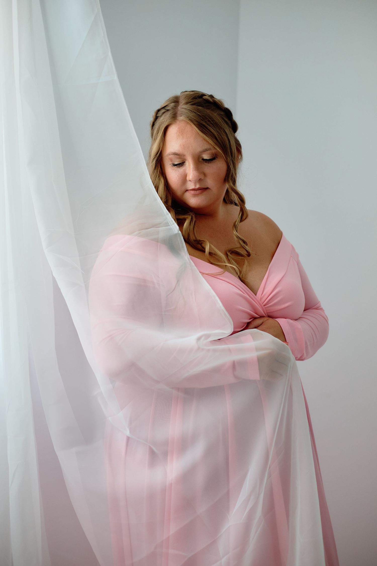 Woman in soft pink wrap dress poses elegantly against white sheer curtains in an intimate boudoir portrait.