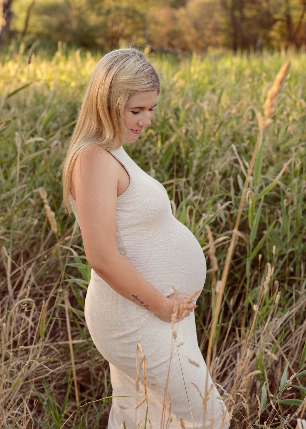 A maternity photoshoot in a golden field shows a woman in white dress cradling her baby bump during sunset.