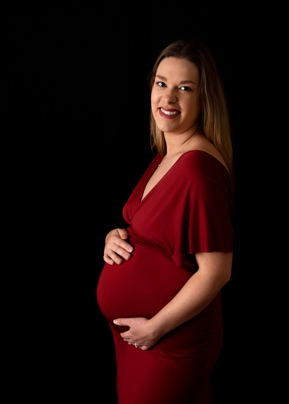 Expectant mother in a deep red dress poses against dark background for maternity portrait.