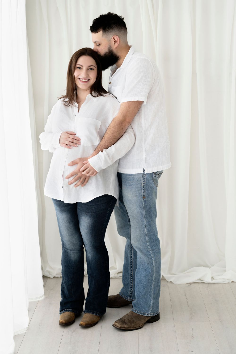 Pregnant woman in white shirt and jeans smiles as her partner embraces her from behind in a bright white studio.